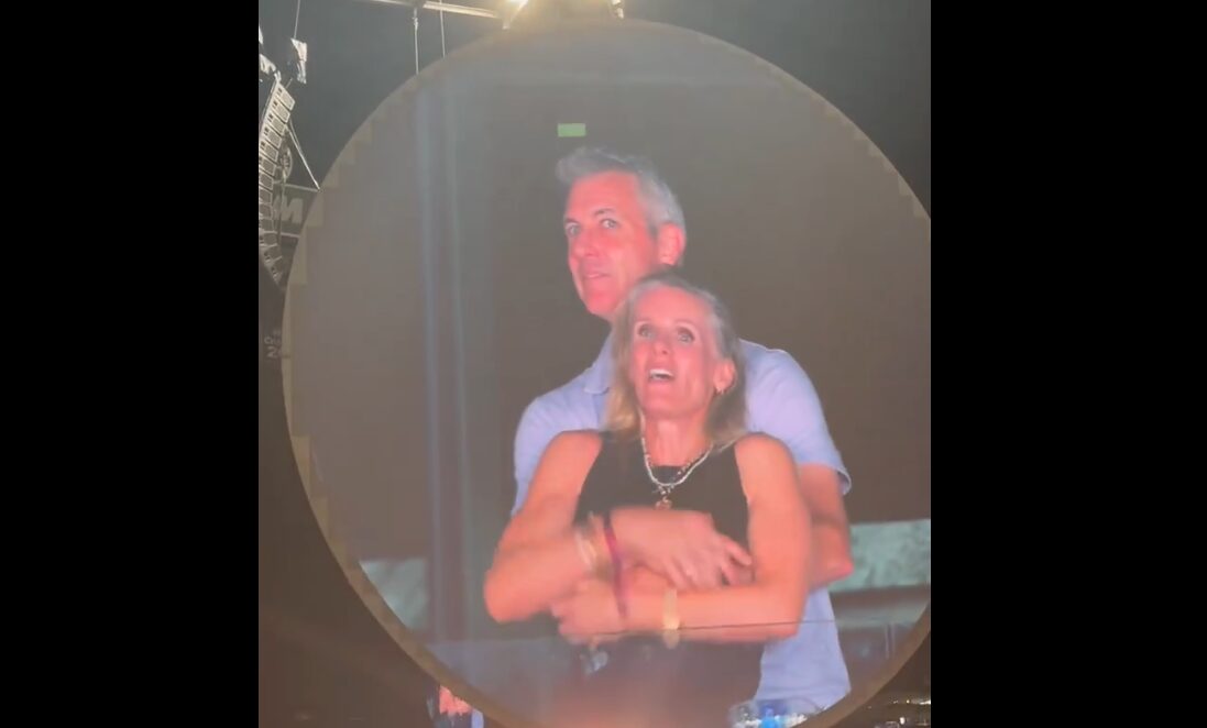 A jumbotron screen at a Coldplay concert showing two people in an awkward embrace, with one person ducking and the other covering their face.