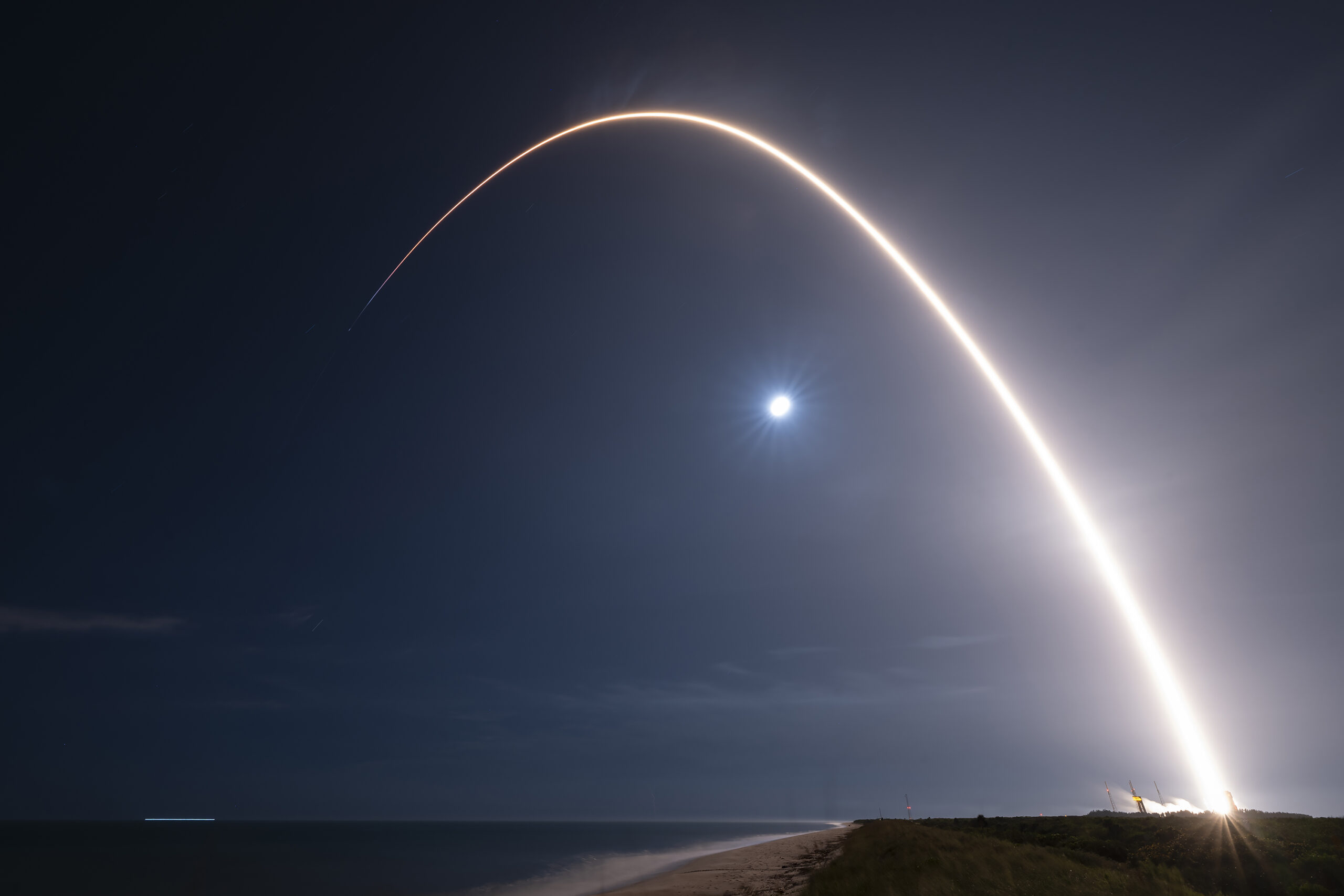 Long exposure photo of a SpaceX Falcon 9 rocket launching at night from Cape Canaveral, with a bright arc of light tracing its ascent against a dark sky and the moon visible.