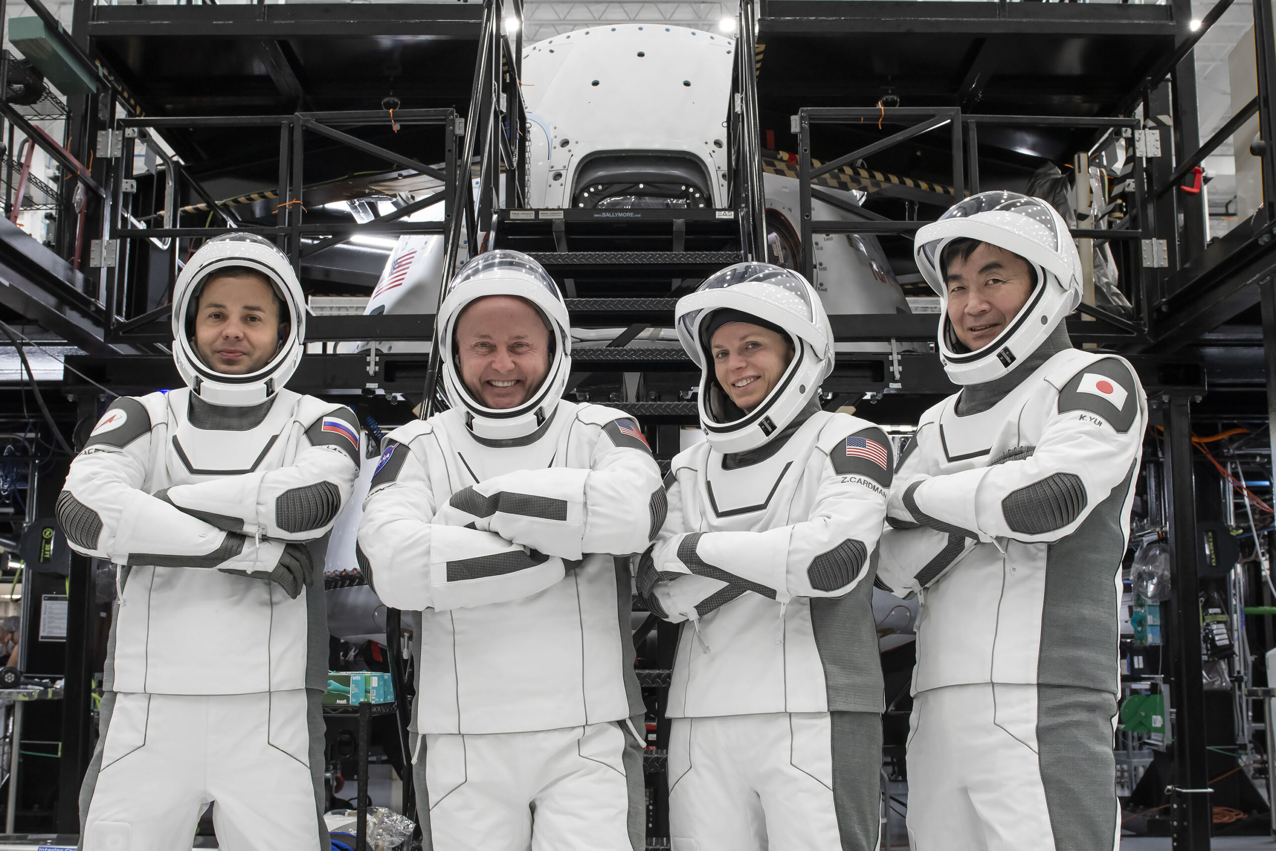 Four astronauts in white SpaceX spacesuits, including one Japanese astronaut, smiling and standing in a spacecraft processing facility with hardware in the background.