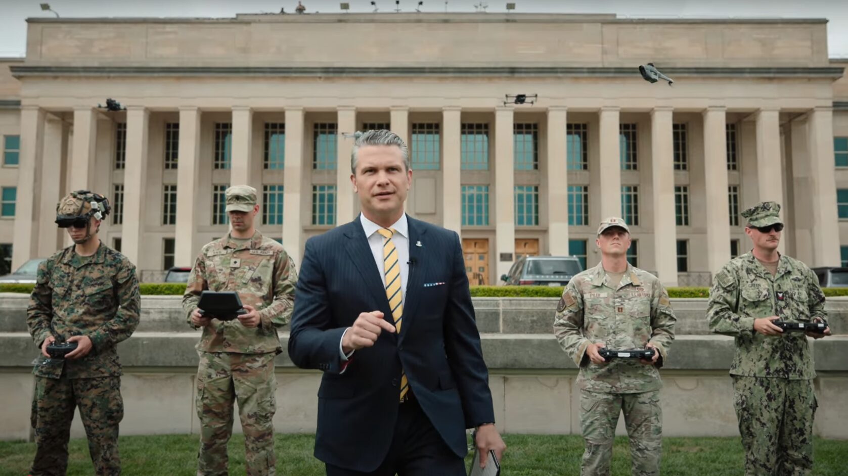 Pete Hegseth, in a suit, stands in front of the Pentagon with four service members holding drone controllers, one FPV drone flies above.